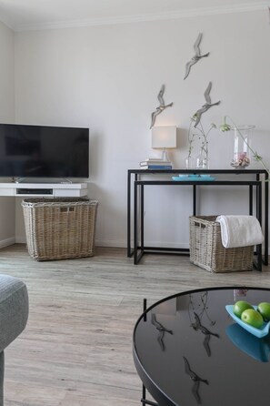 Un salon doté d’un téléviseur à écran plat, d’une table agrémentée d’objets décoratifs, de paniers en osier et d’une étagère murale où sont posés des livres et une lampe.