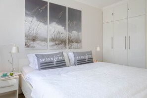 A bedroom with a bed, white walls, a large painting, and a wardrobe. - Meeresrauschen - Haus Dünenburg, Elisabethstraße 9 - Diese Freundlich Eingerichtete 2-zimmer-wohnung (Westerland)