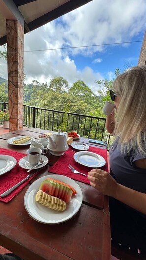 A person seated at a wooden table with a red tablecloth, enjoying a meal with a view of lush greenery and a partly cloudy sky. - HERITAZ VILLA ELLA (Ella)