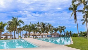 A resort pool area with a clear view of the ocean, surrounded by palm trees and thatched-roof structures. - NEW Beachfront Marítima Condos 2 Pools (Nuevo Nayarit)