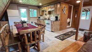 Interior rústico de una cabaña con una mesa de comedor de madera, una cocina con armarios de madera y una zona de estar acogedora con un sofá de cuero marrón.