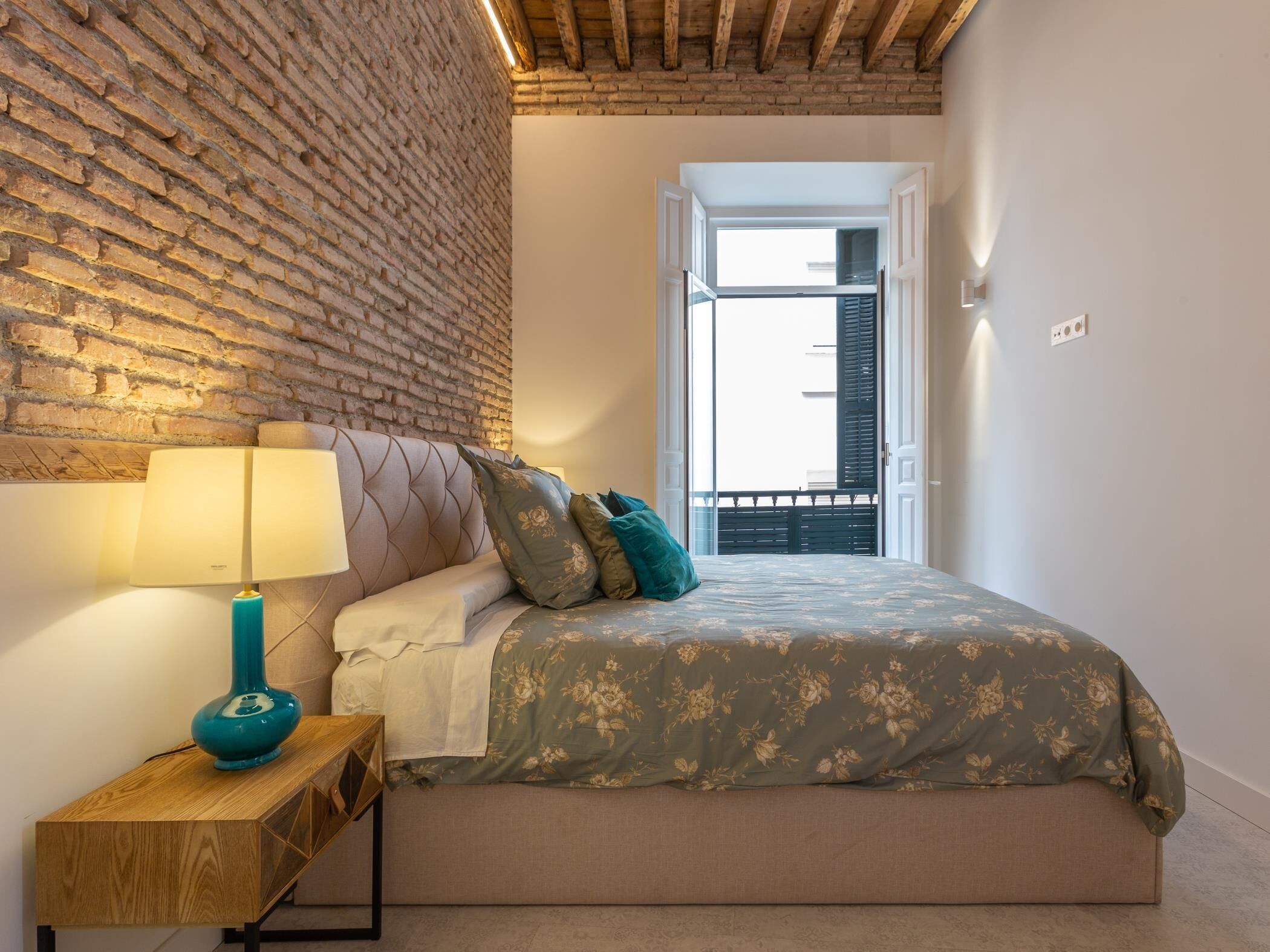 Un dormitorio con pared de ladrillo, cama con ropa de cama floral, una lámpara color turquesa y una mesita de noche de madera.