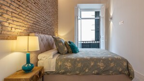 Ein Schlafzimmer mit einer Ziegelwand, einem Bett mit floralem Bettzeug, einer türkisfarbenen Lampe und einem Holfnachtkästchen.