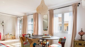 A dining area with a round table, colorful chairs, a hanging light fixture, and a view of buildings through large windows. - Central and bright apartment with balconies (Málaga)