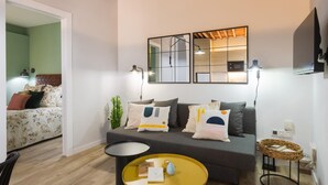 Un salon moderne avec un canapé gris, une table basse jaune et une table d’appoint noire. On y trouve deux lampes fixées au mur et un téléviseur à écran plat.