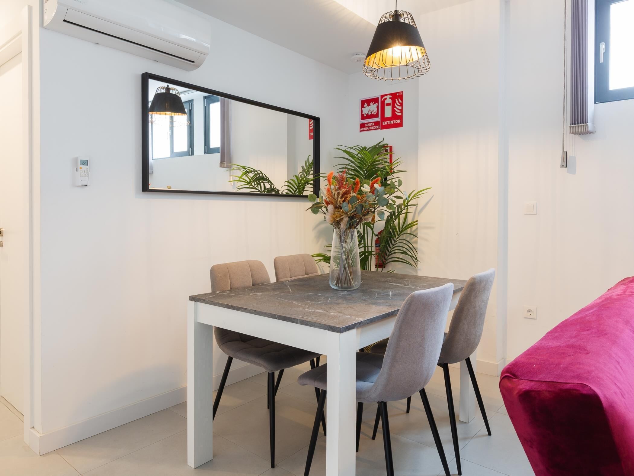 Un comedor con una mesa blanca, sillas grises, un jarrón con flores y una lámpara colgante.