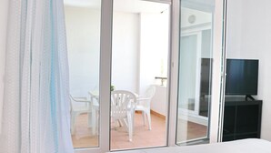 A hotel room with a bed, a TV, and a sliding glass door leading to a balcony with white chairs and a small table. - Apartamento Bateles Cerca de la Playa (Conil de la Fra.)