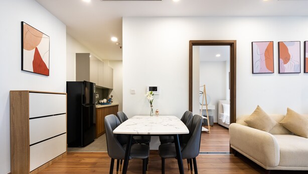 A modern living room with a beige sofa, a dining table with six chairs, and a kitchen area with a refrigerator and cabinets. - Eco Green Apartment- Minimalist Home (Ho Chi Minh City)