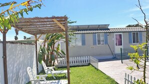 A patio area with a pergola, two white chairs, a small garden, and a building with a tiled roof and windows. - Bungalow Puntalejo in Conil (Conil de la Frontera)