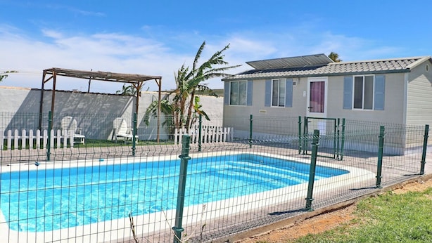 A swimming pool with a fenced deck, a covered seating area, and a small building with windows and shutters. - Bungalow Puntalejo in Conil (Conil de la Frontera)