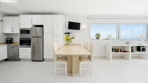 Una cucina moderna con mobili bianchi, un'area pranzo con tavolo e sedie, una TV a parete e una finestra con vista su palme.