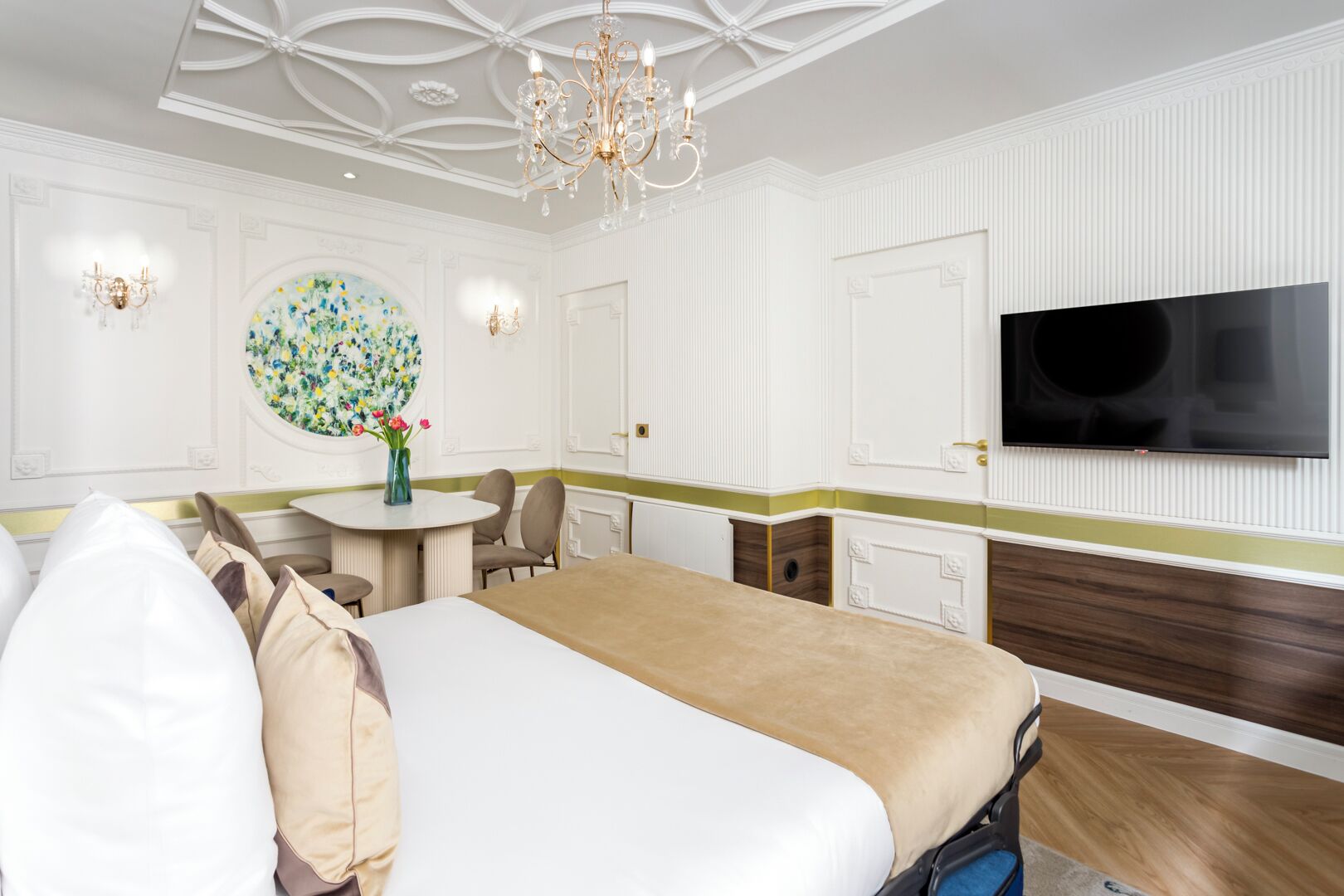 Ett hotellrum med en stor säng, ett runt bord med stolar, en kristallkrona, en tv som är monterad på väggen och ett blommotiv på väggen.