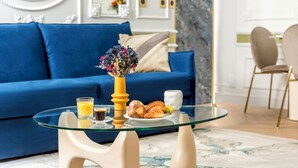 Ein modernes Wohnzimmer mit einem blauen Sofa, einem Glastisch mit Frühstücksangebot und einer dekorativen Vase.