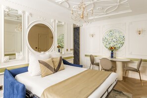 Ein Hotelzimmer mit einem großen Bett, einem runden Spiegel, einer Kristallleuchtre, einem Essbereich und einem runden Tisch mit Blumen.