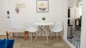 Ein moderner, minimalistischer Essbereich mit einem weißen Tisch, zwei weißen Stühlen, einer kleinen Topfpflanze und einem gerahmten Kunstwerk an der Wand.