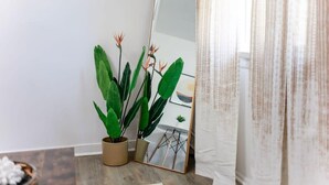 Een kamer met een grote spiegel die een plant en een stoel weerspiegelt. Op de vloer staat een potplant en het gordijn is gedeeltelijk open.