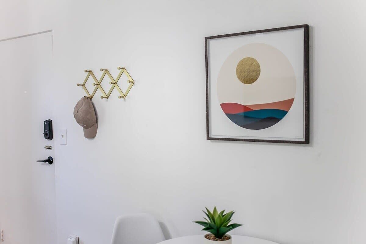 A minimalist room with a white door, a framed abstract painting, a geometric wall decoration, and a small potted plant on a table.