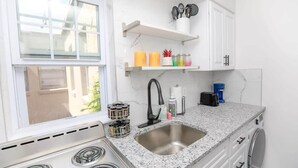 A kitchen with a stainless steel sink, white cabinets, a window, and a countertop with various items. - Anchor Breeze | Sojourn | Steps to the Beach (Norfolk)