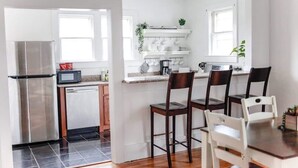 A compact kitchen with a refrigerator, microwave, and sink. There are white cabinets, a countertop with a plant, and a dining area with chairs and a table. - Sojourn | Hideaway Bay | 2 BR (Norfolk)