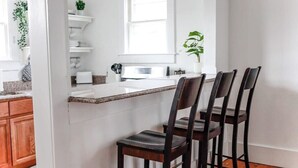 A kitchen with a white countertop, wooden chairs, and a window. - Sojourn | Hideaway Bay | 2 BR (Norfolk)