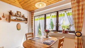 Ein Holzespeisesaal mit Stühlen, ein Fenster mit Vorhängen, eine Heizung und Regale an der Wand mit verschiedenen Gegenständen.