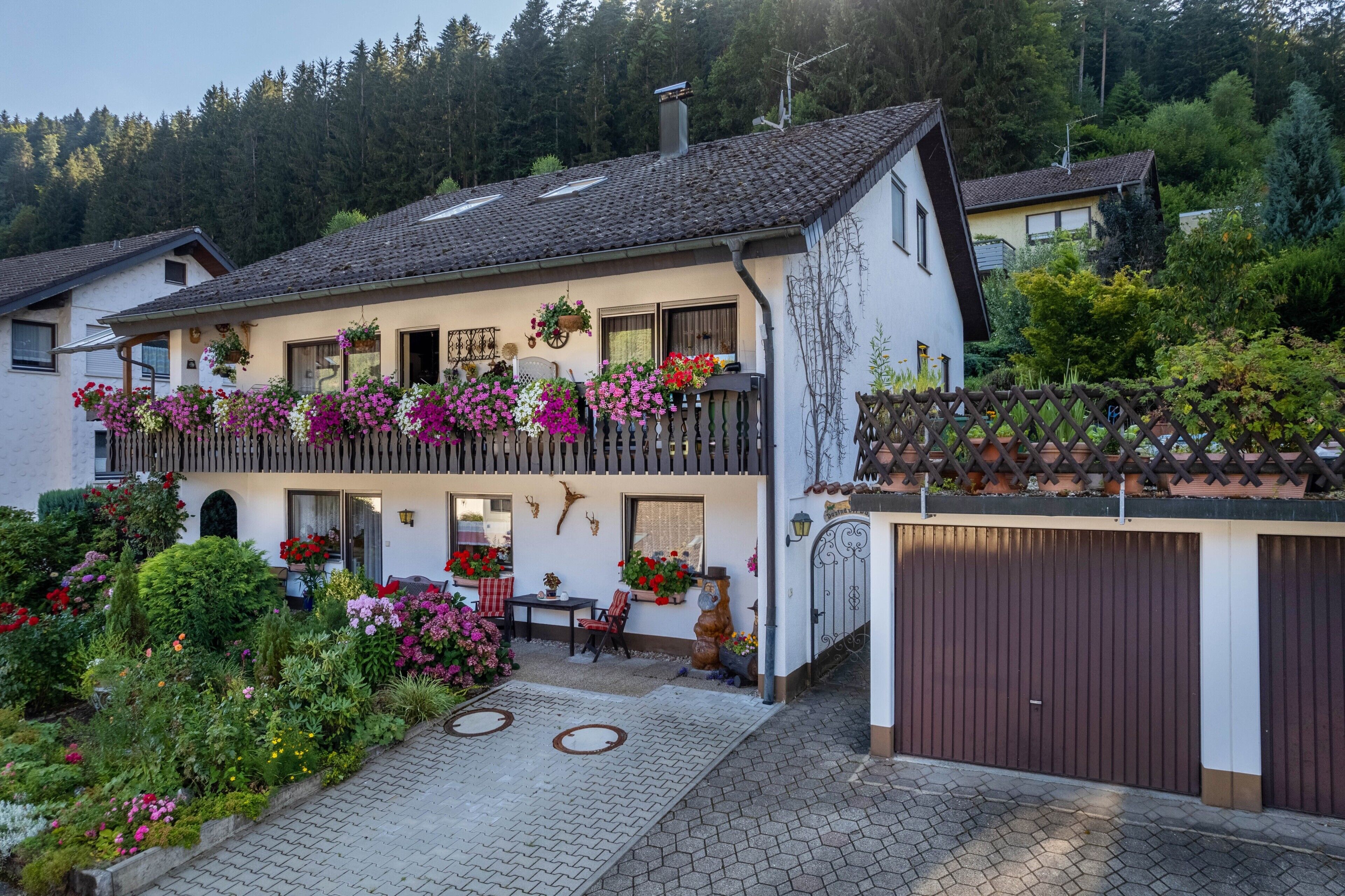 Ein zweigeschossiges Haus mit Balkon, Garage und Garten mit verschiedenen Pflanzen und Blumen.