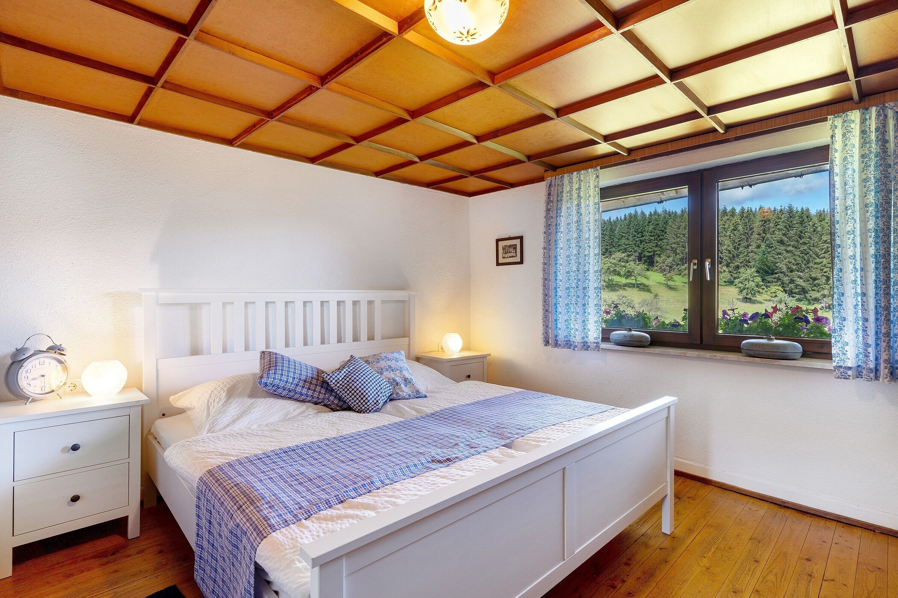Ein Schlafzimmer mit Holzboden, einem Bett mit weißem Kopfteil, zwei Nachttischen mit Lampen und einem Fenster mit Blick auf einen Wald.