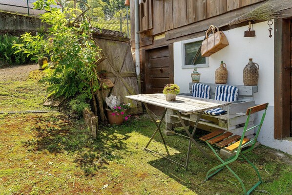 A rustic outdoor seating area with a wooden table and green metal chair, a potted plant, and wicker baskets on a white wall.