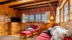 A wooden cabin interior with a sofa, a chair, a television, and a window with curtains. - Holiday Home 'Blockhaus Paepke' with Private Terrace and Private Garden (Mühlenbach)