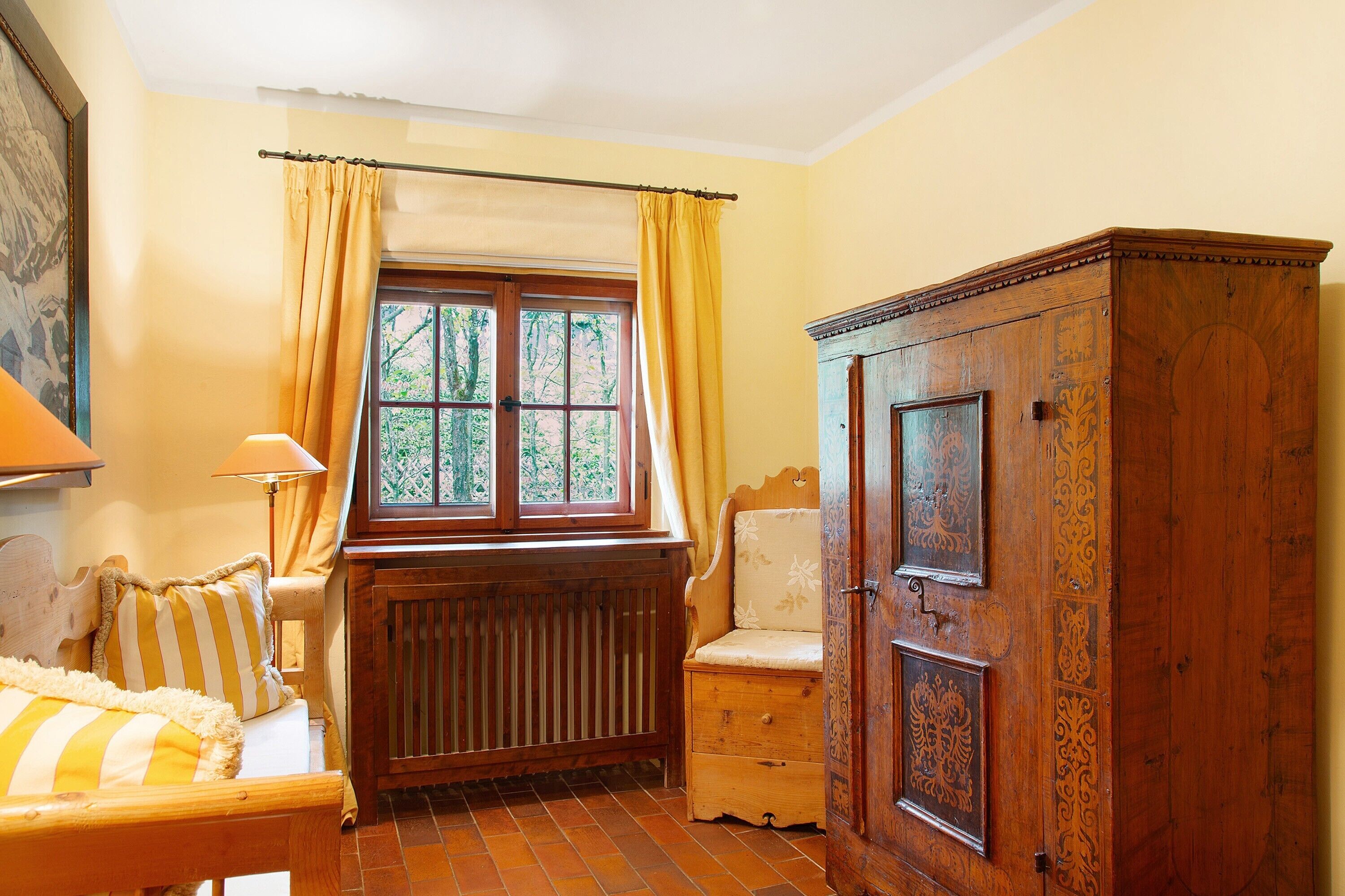 Quarto com radiador de madeira, janela com cortinas amarelas, armário de madeira e cama com roupa de cama listrada.