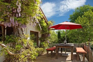 Een houten terras met een tafel en stoelen, een rode parasol, en een huis met klimplanten.