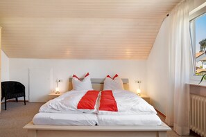 A bedroom with a wooden ceiling, a bed with white and red bedding, bedside lamps, a black chair, and a window with curtains. - Apartment 'Ferienwohnung Lange' with Mountain View, Balcony and Wi-Fi (Waldstetten)