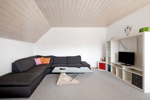 A modern living room with a sectional sofa, a glass coffee table, a television on a white entertainment unit, and a sloped ceiling. - Apartment 'Ferienwohnung Lange' with Mountain View, Balcony and Wi-Fi (Waldstetten)