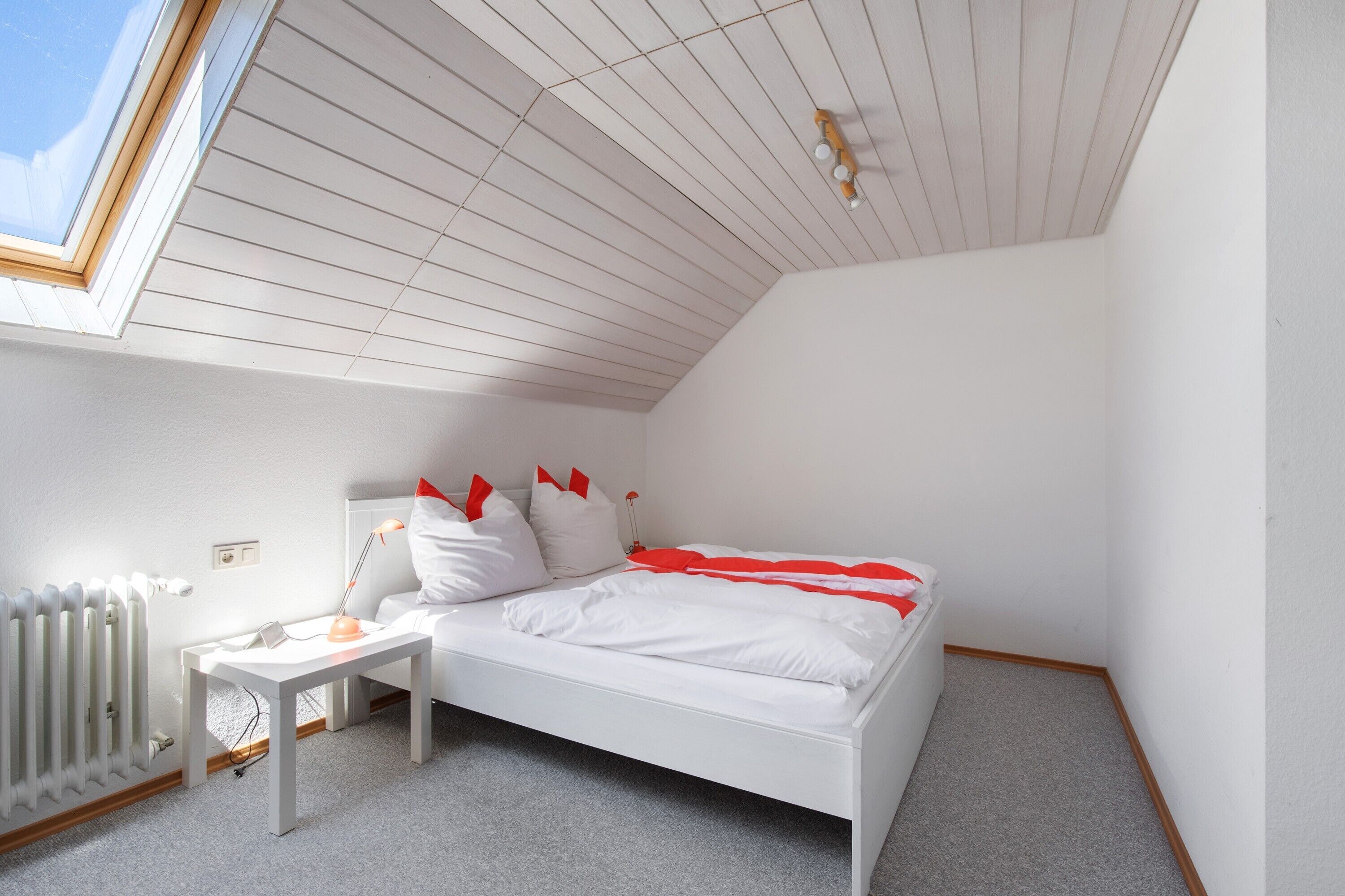 A bedroom with a sloped ceiling, a skylight, a white bed with red pillows, a bedside table with a lamp, and a radiator.