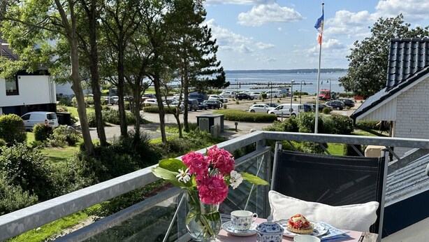 Un balcón con una mesa dispuesta para tomar el té, un jarrón con flores y vistas al mar y coches estacionados.