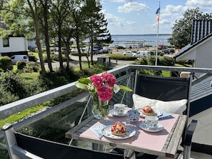 A balcony with a table set for tea, a vase with flowers, and a view of the sea and parked cars.