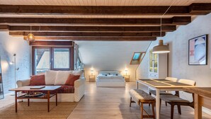Ein modernes Wohnzimmer mit Sofa, Couchtisch und Esstisch. Im Hintergrund ist ein Bett, ein Fenster und eine Decke mit sichtbaren Balken zu sehen.