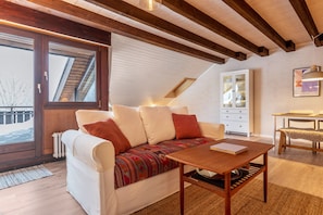 Une chambre à coucher confortable, aménagée en mezzanine avec un plafond en pente, un canapé agrémenté de coussins à motifs, une table en bois et une commode blanche.