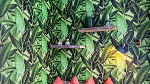 Une pièce avec un mur orné d’un motif luxuriant de feuilles vertes, deux étagères en bois sur lesquelles sont posées des figurines, une lampe suspendue et un lit en bois avec des oreillers rouges et blancs.