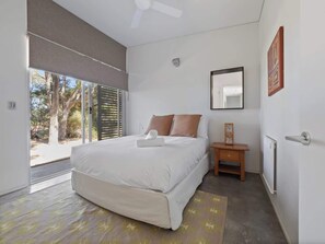 A modern bedroom with a large bed, a small bedside table, a mirror, and a view of the outdoors.