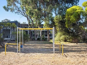 A playground with yellow and white equipment, surrounded by trees and a building in the background.