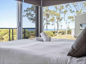 Una cama con sábanas blancas y una toalla enrollada, un balcón con vistas a árboles y un edificio, y un cielo despejado.