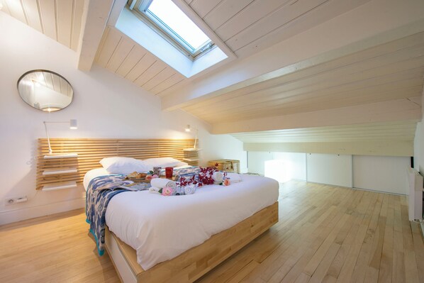 Ein modernes Schlafzimmer mit Oberlicht, Holzboden und einem ordentlich bezogenen Bett mit Handtüchern und Blumen.