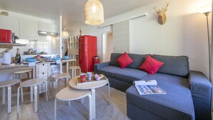 A compact living space with a kitchenette, a grey sofa with red cushions, a dining table, and a ceiling-mounted light fixture.