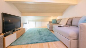 A modern living room with a flat-screen TV on a wooden stand, a beige sofa with cushions, and a teal rug on wooden flooring.
