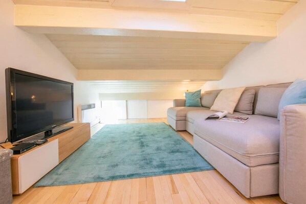 Uma sala de estar moderna com uma TV de tela plana sobre um suporte de madeira, um sofá bege com almofadas e um tapete verde-água sobre piso de madeira.