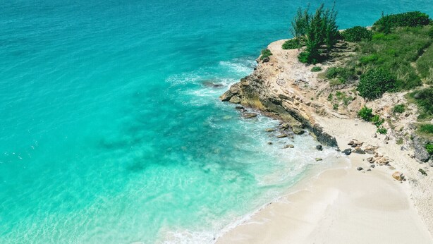A beach with clear turquoise water, a sandy shore, and a rocky outcrop with vegetation.