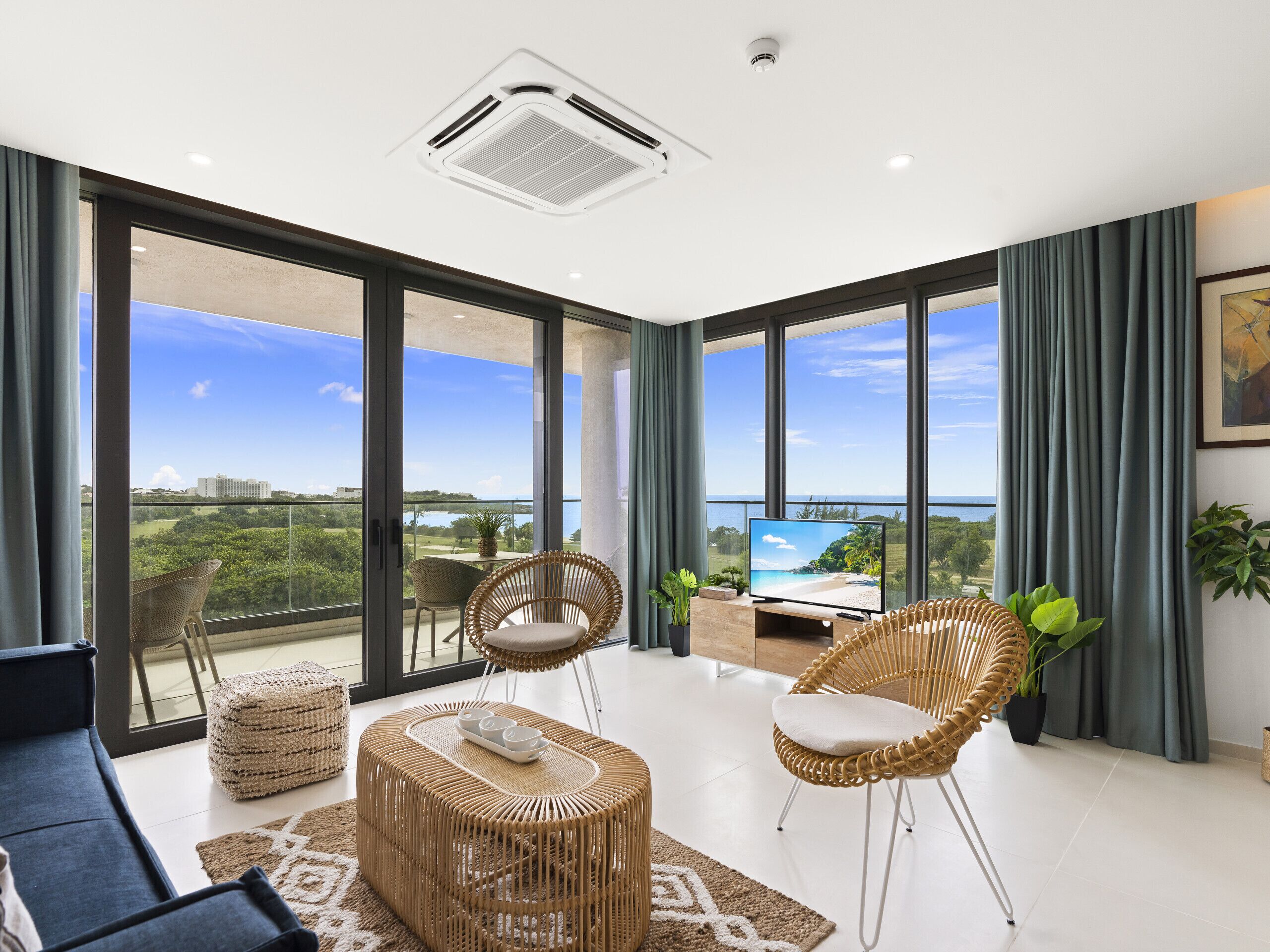 A spacious living room with large windows, a flat-screen TV, wicker furniture, and a view of a beach and cityscape.
