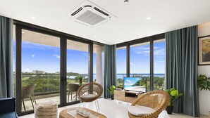 A spacious living room with large windows, a flat-screen TV, wicker furniture, and a view of a beach and cityscape.
