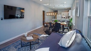 A modern living room with a sofa, a coffee table, and a TV. There is a dining area with a table and chairs. The kitchen is visible in the background.